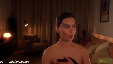 Media: Video of a topless woman with dark hair, wearing black gloves, standing in a dimly lit living room with beige curtains and a modern sofa.