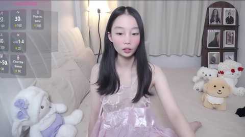 A video of an Asian woman with long black hair in a pink dress, surrounded by stuffed animals, in a cozy, softly lit living room.