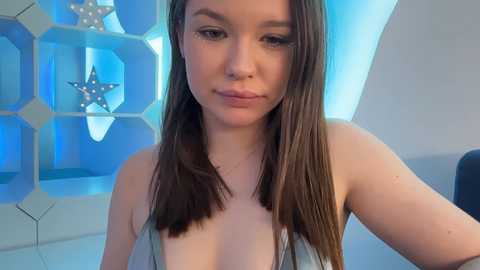 Video of a young Caucasian woman with long brown hair, wearing a low-cut sleeveless top, posing indoors with a blue geometric wall and star-shaped decor in the background.