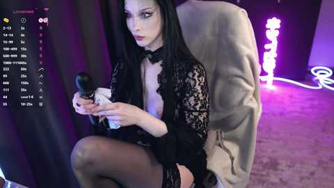 Video of a pale-skinned woman with long black hair and dark makeup, wearing a black lace top and stockings, holding a black microphone. She sits in a dimly lit room with purple and white lights, behind a DJ booth.