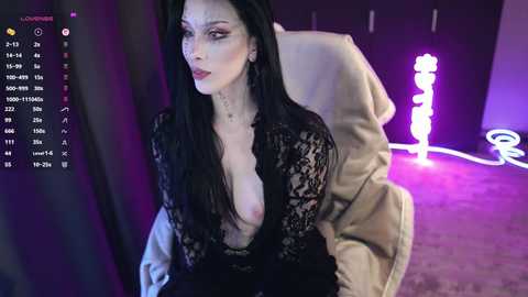 Video of a pale-skinned woman with long black hair, wearing a black lace dress exposing her breasts, sitting on a bed in a dimly lit room with purple lighting.
