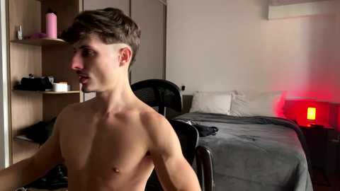 Video of a shirtless young man with short brown hair, standing in a dimly-lit bedroom. The background includes a bed, a red lamp, and a chair.