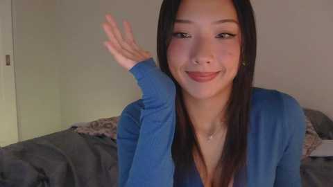 Video of an East Asian woman with long black hair, light skin, and almond-shaped eyes, smiling, waving with one hand, wearing a blue cardigan, indoors with gray bedspread in background.