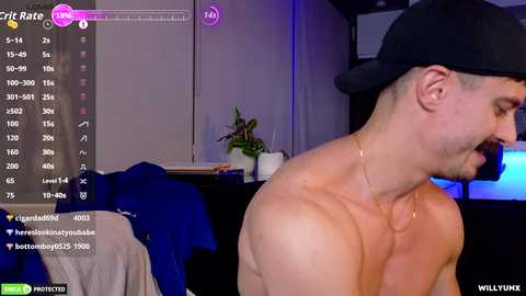 Video of a shirtless, fit man with a black cap, smiling, in a dimly lit room with a blue screen and potted plants.