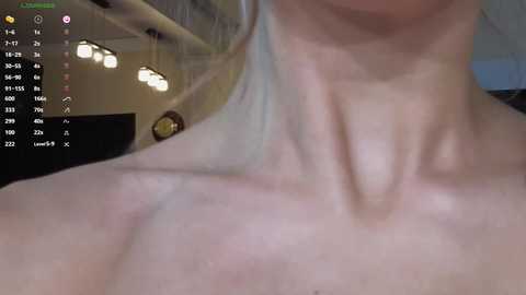 Media: A close-up video of a woman's bare upper torso, showcasing her collarbone and neck. The background features a dimly lit room with hanging lights and a clock. The image is slightly blurry.