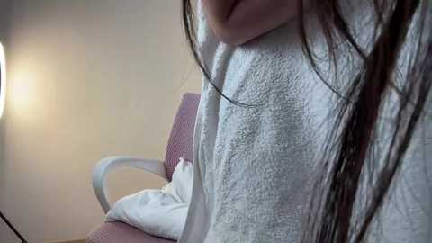 Video of a woman with long, wet, dark hair, wearing a white towel, sitting on a pink chair in a dimly lit room.