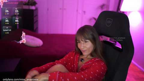 Video of a woman in a red patterned pajama top, sitting in a black gaming chair with a white pillow, in a dimly lit bedroom with purple lighting.