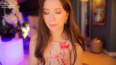 Video of a young woman with long brown hair, wearing a floral sleeveless top, standing indoors with blurred background of flowers and framed artwork.