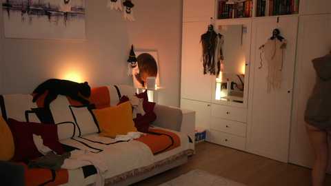 Media: Video of a cozy, dimly lit bedroom with a white bed adorned with orange, red, and white pillows. A black cat stands on the bed, and a person wearing a grey dress is partially visible.