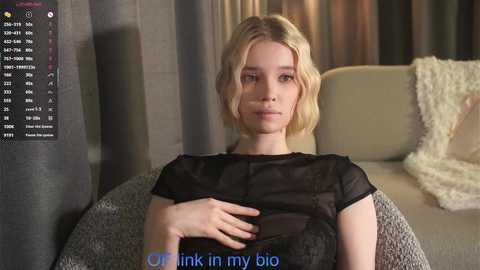 Media: A video of a young woman with short blonde hair, wearing a sheer black top, sitting on a beige sofa with fluffy pillows. She has a neutral expression and the text \"Or think in my bio\" is overlaid.