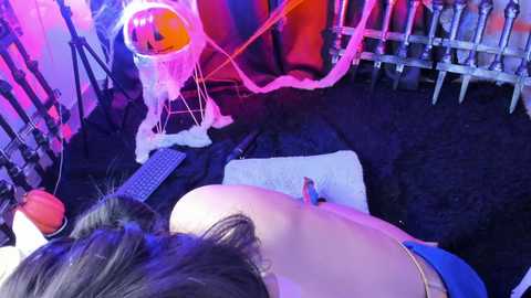 Media: Video of a woman with dark hair, topless, lying on her stomach on a white pillow, in a room with a black floor, purple lighting, and a white bucket with a red rope.
