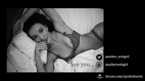 Black-and-white video of a slender, light-skinned woman with dark hair, lying on a bed in lingerie, gazing seductively at the camera.