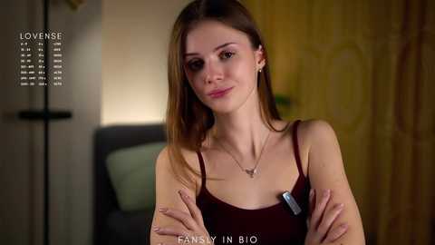 Video of a young Caucasian woman with light skin, straight brown hair, wearing a sleeveless dark top, standing indoors with arms crossed, smiling, background includes a wooden door and blurred furniture.