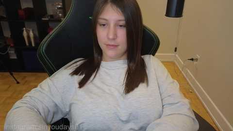 Video of a young Caucasian woman with long brown hair, wearing a light gray sweater, sitting in a black gaming chair with green accents, in a modern, sparsely decorated room with a wooden floor.