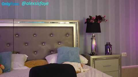 Video of a chic, modern bedroom with a tufted gray headboard, white bedding, light blue pillows, and a matching nightstand with a black lamp and floral arrangement. The wallpaper features vertical stripes in soft hues.