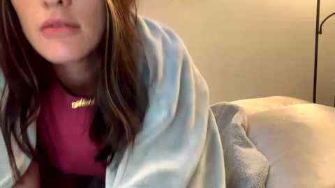 Video of a woman with fair skin, light brown hair, and light makeup, wearing a red shirt, wrapped in a light blue blanket, lying on a bed with a beige blanket and white pillow.