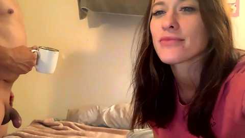 Video of a fair-skinned woman with long brown hair and a red shirt, sitting on a bed with a beige blanket, holding a white mug. A partially visible man stands on the left.