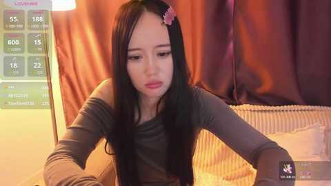 A video of a young Asian woman with long black hair, wearing a gray shirt, seated at a desk, in a dimly lit room.