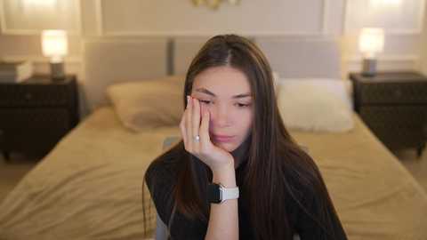 Media: A video of a young woman with long brown hair, wearing a black sweater and a smartwatch, resting her head on her hand in a dimly lit bedroom with beige linens and two bedside lamps.