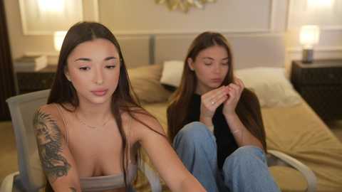 Media: Video of two young women with long dark hair, one with a tattoo sleeve, sitting on a bed in a cozy, dimly lit bedroom.
