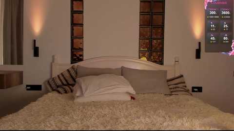 Video of a cozy, modern bedroom with a plush cream shag rug, white bed, beige pillows, white walls, and two black wall sconces flanking a window with a view of a city skyline.