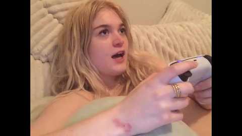 Media: Video of a young woman with long, messy blonde hair, fair skin, and light makeup, playing a video game on a white controller, lying on a textured beige pillow.