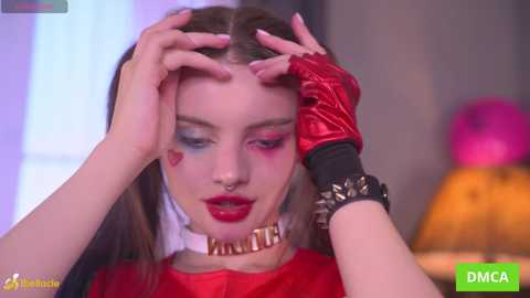 Video of a fair-skinned woman with long brown hair, wearing red lipstick and multicolored eye makeup. She's in a red top and black fingerless gloves, with a spiked bracelet, in a dimly lit room.