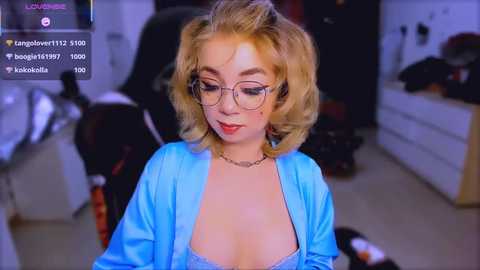 Video of a blonde woman with glasses, wearing a blue robe, standing in a dimly lit room with a white dresser and a bicycle in the background.