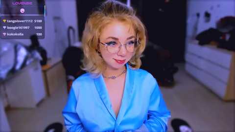 Video of a young blonde woman with shoulder-length hair, wearing a blue blouse, glasses, and a necklace, standing in a dimly lit kitchen.
