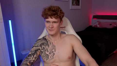 Video of a shirtless young man with curly red hair and a detailed black tattoo sleeve, sitting on a white gaming chair in a dimly lit bedroom with pink and blue lighting, a bed, and framed pictures on the wall.