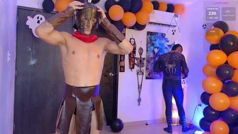 Video of a Halloween party scene with a muscular, shirtless man wearing a mask and leather vest, surrounded by orange and black balloons, skeletons, and a man in a spider suit.