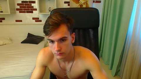 Video of a shirtless young man with short brown hair, seated on a black office chair in a modern bedroom with white brick walls, green curtains, and a white bed.