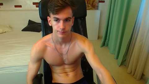 Video of a shirtless, slender young man with short brown hair, sitting in a black gaming chair in a modern bedroom with white walls, green curtains, and a bed.