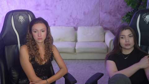 Media: Video of two young women in gaming chairs, one in a black lace top, the other in a black shirt, in a dimly lit room with a white sofa and purple wall.