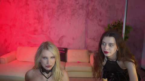 Media: Video of two women in a dimly lit room with pink and red lighting. The blonde woman in the foreground has pale skin and wears a black choker. The brunette woman in the background has long hair and is dressed in a black top.