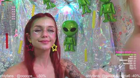 Video of a topless, pale-skinned woman with long red hair and a star tattoo on her face, smiling, surrounded by green alien toys and shiny mylar.