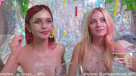 Video of two young women with fair skin and long hair, one with red, the other blonde, wearing star-shaped glitter on their faces, sitting side-by-side in front of a shiny, colorful background. Text overlays indicate OnlyFans and content details.