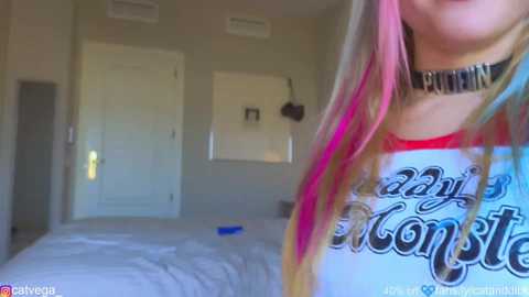 Media: Video of a woman with long, multicolored hair and a choker, wearing a white \"The Last Starfighter\" shirt, standing in a minimalist bedroom with a bed and closed doors.