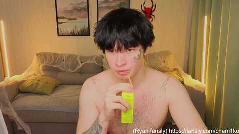 Video of a shirtless, slender, fair-skinned, black-haired young man holding a bright yellow straw. He sits on a beige couch in a cozy living room with a painting and red antlers in the background.