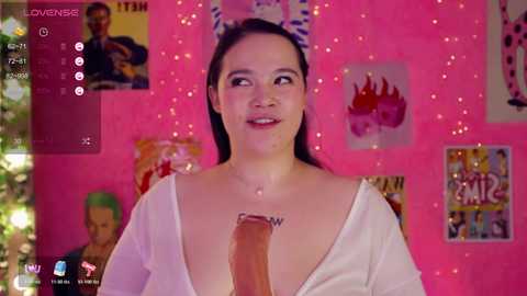 A video of a young Asian woman with long black hair, wearing a white shirt, with an erect penis inserted into her mouth. The background features vibrant pink walls with posters and Christmas decorations.