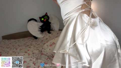 Media: Video of a woman in a white, backless wedding dress with intricate lace-up details, standing beside a black cat with green eyes, on a floral-patterned carpet in a dimly lit room.