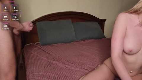 A video of a nude man and woman in a bedroom, standing next to a bed with green pillows and a red blanket.