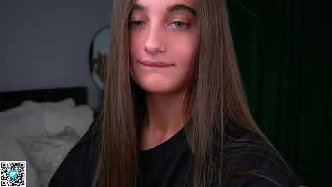 Media: Video of a young person with long, straight brown hair, wearing a black shirt, standing indoors near a bed with white sheets, dark green curtains, and a blurred circular mirror in the background.