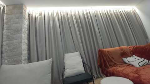 Video of a cozy, modern living room with a gray curtain backdrop, a plush, orange sofa, and a white chair; warm, ambient lighting from above.