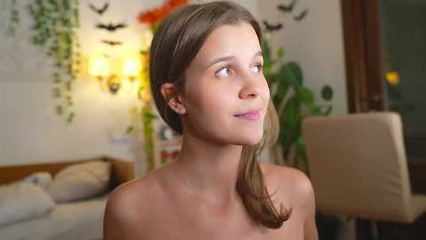 Video of a fair-skinned young woman with light brown hair, smiling, topless, standing indoors. Background features a beige sofa, green plants, and a warm light.