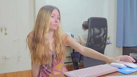 Video of a young woman with long, wavy blonde hair, wearing a sleeveless, patterned dress, typing on a pink keyboard in an office with a desk, chair, and blue curtain.