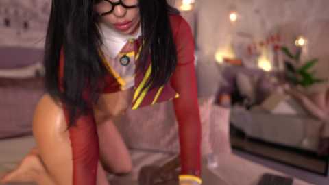 Video of a woman with long black hair and glasses, wearing a red crop top and yellow tie, kneeling on a bed in a dimly lit, cozy bedroom with a mirror and potted plants.