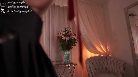 Media: Video of a dimly lit, elegantly decorated room with a black figure in the foreground, a white tufted chair, a glass table with a turquoise vase of pink flowers, and sheer curtains.