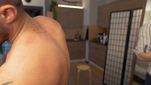 Video of a shirtless man with a tattooed back, red sunburn, and visible shoulder blades. He stands in a modern kitchen with wooden cabinets, a black-and-white screen, and a stool.