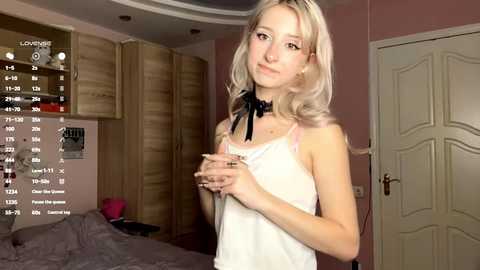 Video of a young, slim, fair-skinned, blonde woman in a white tank top, standing in a pink-painted bedroom with a closed door, a bed, and a wooden closet in the background.